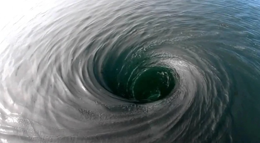 A whirlpool in open water, seen from above: water spiralling inward toward a dark opening at the centre.